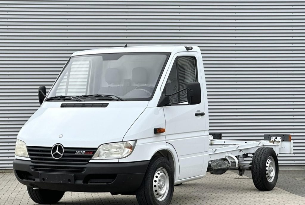 White Mercedes-Benz Sprinter cab chassis seen from the front/right, with no cargo body attached, parked on a paved lot against a corrugated metal wall.
