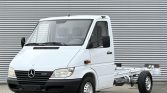 White Mercedes-Benz Sprinter cab chassis seen from the front/right, with no cargo body attached, parked on a paved lot against a corrugated metal wall.