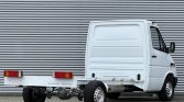 White Mercedes-Benz Sprinter cab with empty flatbed chassis, showing rear lights and exposed drivetrain against a corrugated metal wall.