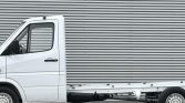 White cab-over truck with an empty flatbed parked in front of a corrugated metal wall.