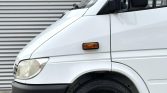 White cargo van shown from the front-left side, parked against a corrugated metal wall, featuring a round headlight and side mirror.