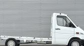 White cab-forward truck with an empty flatbed frame parked beside a corrugated metal wall.