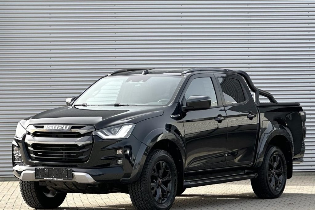 Black Isuzu pickup truck with a bold chrome grille, large tires, and a tubular roll bar parked in front of a corrugated metal wall.