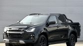 Black Isuzu pickup truck with a bold chrome grille, large tires, and a tubular roll bar parked in front of a corrugated metal wall.