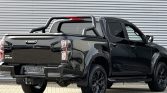 Black Isuzu D-Max pickup parked in front of a corrugated metal wall, rear three-quarter view showing cargo bed and roll bar.