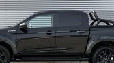 Side view of a black four-door pickup truck with a bed roll bar, parked against a corrugated metal wall.”] ,