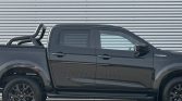 Side profile of a black double-cab pickup truck with a roll bar in the bed, parked against a corrugated metal wall.