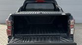 Open-flat bed of a black pickup truck seen from the rear, showing a empty bed with a ribbed liner and roll bar behind the cab in front of a corrugated metal wall.