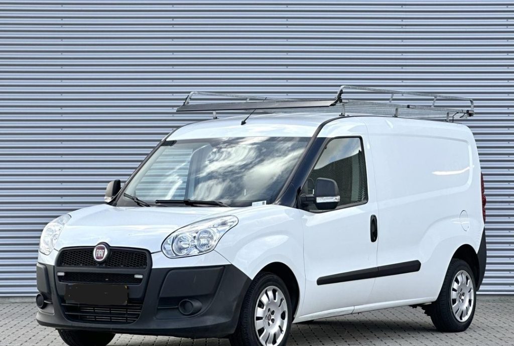 Front three-quarter view of a white Fiat commercial cargo van with a roof ladder rack, parked on a paved surface against a corrugated metal wall.