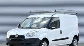 Front three-quarter view of a white Fiat commercial cargo van with a roof ladder rack, parked on a paved surface against a corrugated metal wall.