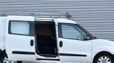 White cargo van with side sliding door open, roof ladder rack, parked against a corrugated metal wall.