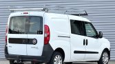 Rear and side view of a white Fiat Doblo Maxi cargo van with a roof ladder rack, parked in front of a corrugated metal backdrop.