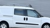 White compact van with a roof rack parked in front of a corrugated metal wall; side sliding door and black bumper details visible.