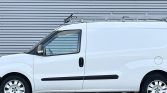 Side view of a white cargo van with a roof ladder rack parked in front of a corrugated metal wall