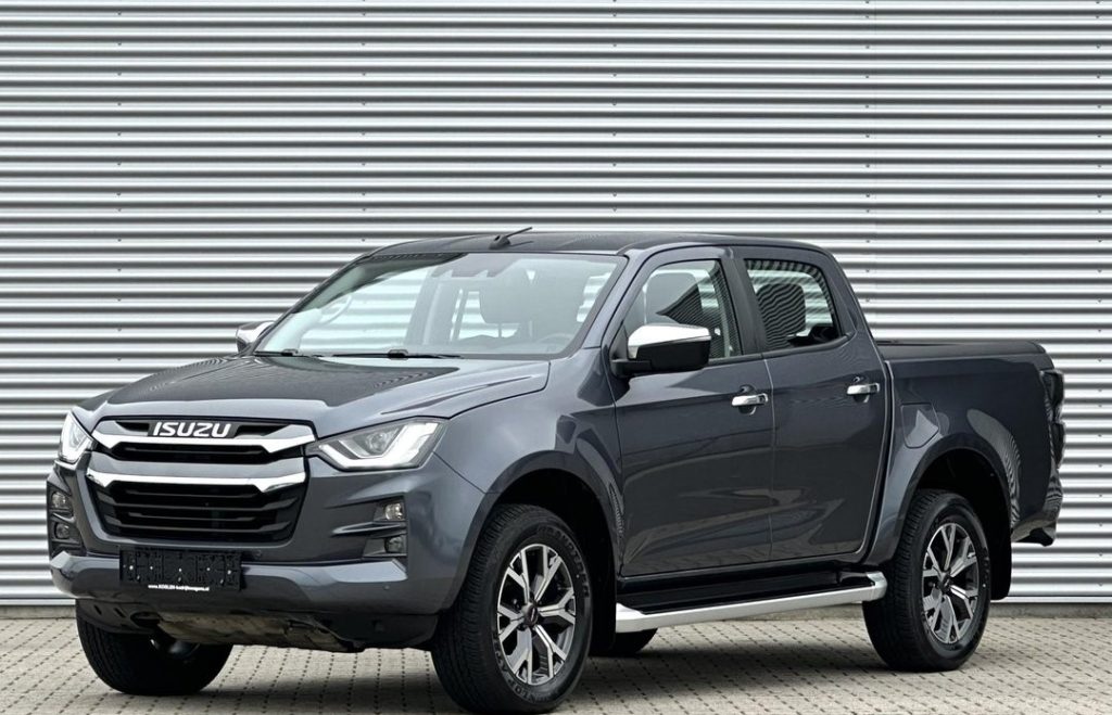 Grey Isuzu pickup truck with crew cab, parked on a paved surface in front of a corrugated metal wall.