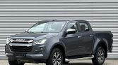 Grey Isuzu pickup truck with crew cab, parked on a paved surface in front of a corrugated metal wall.