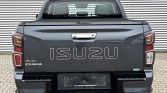 Rear view of a gray Isuzu D-Max pickup truck parked on a brick surface, prominent ISUZU lettering on the tailgate.