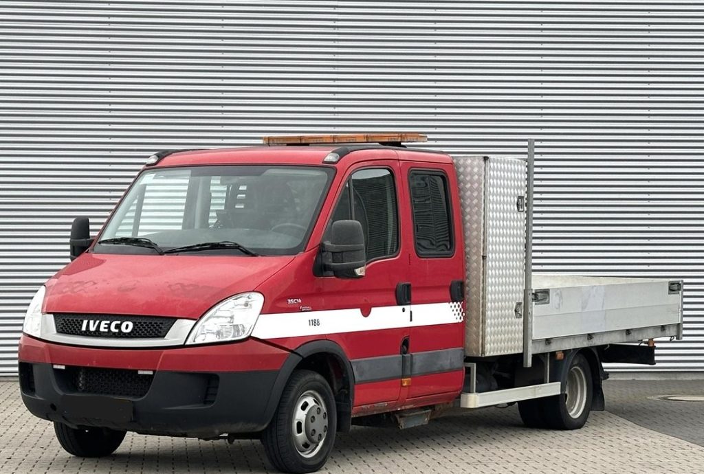 Red Iveco utility truck with a flatbed and diamond-plate side panel, parked in front of a corrugated metal wall.