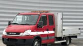 Red Iveco utility truck with a flatbed and diamond-plate side panel, parked in front of a corrugated metal wall.