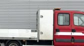 Red utility van with a flatbed cargo area, side view, parked against a metal building with a white stripe along the door area.