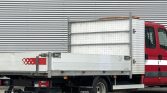 Red utility truck with a flatbed and diamond-plate storage box, parked against a corrugated metal wall, seen from the rear side