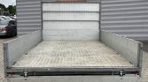 Rear view of an empty metal flatbed trailer with low side rails and a diamond-plate tailgate, parked in front of a corrugated wall.