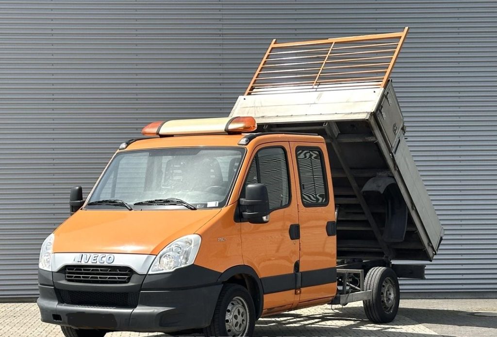 Orange Iveco dump truck with its bed raised, ready to unload, parked in front of a corrugated metal wall.