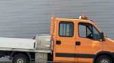 Orange utility truck with a flatbed and metal storage box, parked against a gray corrugated metal wall, with a roof beacon light over the cab.