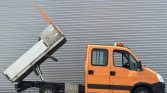 Orange utility truck with a raised dump bed, preparing to unload material in front of a corrugated metal wall.