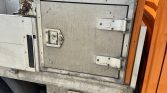 Side view of a weathered metal storage box on a truck, with a lockable hatch and orange cab in the background.