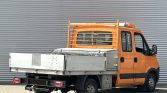 Orange work van with a metal flatbed and toolbox, parked in front of a corrugated metal wall.