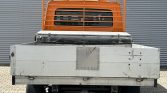 Rear view of a small flatbed truck with an orange protective cab guard and a weathered metal cargo box on a paved surface near a corrugated wall.
