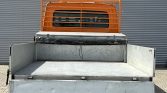 Back of a weathered metal utility trailer with an orange protective cab guard and open cargo bed on a paved surface outside a corrugated wall.