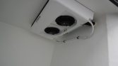 White outdoor air conditioning unit with two circular fans mounted on a white wall/ceiling corner, pipes running along the wall