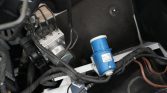Close-up of a blue automotive sensor plugged into a wiring harness in a car's engine bay, with black cables and a silver electronic module nearby