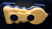 Bright yellow manual release device with two black circular controls and a central wheel, mounted to a dark surface.