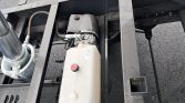 Reservoir for hydraulic fluid with red cap mounted on a metal trailer frame, hoses and a pump nearby
