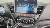 Modern car dashboard with a large central touchscreen showing climate controls, below a row of buttons, and a left-side gear selector dial visible in the foreground.