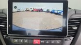 In-car display showing rearview camera feed of a truck yard with parked trucks; Dutch warning text reads 'Let op de omgeving'.