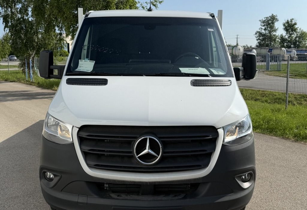 Front view of a white Mercedes-Benz van with a black grille parked outdoors, showing the emblem and headlights facing the camera on a sunny day