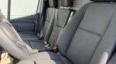 Interior of a gray fabric vehicle front cabin showing the driver seat and a two-passenger bench with visible seat belts and headrests; steering wheel on the left side.