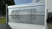 White utility truck with a tall metal cargo gate featuring a grid of square holes in an oval pattern near the back of a building site/yard.