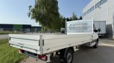 Rear three-quarter view of a white flatbed truck parked in an industrial area beside a large gray warehouse, with another truck in the background.