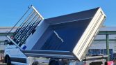 White tipped-dump truck with a raised cargo bed for unloading, exposing a large metallic inner container and perforated side panels on a clear day.