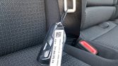 Car key fob with a white tag and QR code hanging from the seat belt latch on a dark fabric car seat near the buckle.