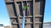 Dump truck bed raised in the air by a hydraulic ram, viewed from underneath; a green circular joint at the base of the ram is visible against the metal structure.