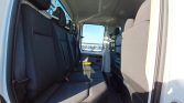 Interior of a white van showing black bench seating along the left, with a window and door visible and sunlight flooding the cabin.