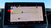 In-car navigation touchscreen showing a map with route markers, streets, and a blue car icon on the right edge in daylight