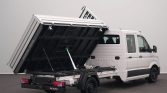 White cargo van with a black tipping dump bed raised to unload material, showing hydraulic struts and rear lights nearby.