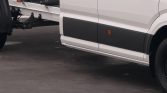 Close-up of a white van with black lower side panel and an attached trailer hitch/framing at the rear, showing part of the wheel and ground.](interpretable)